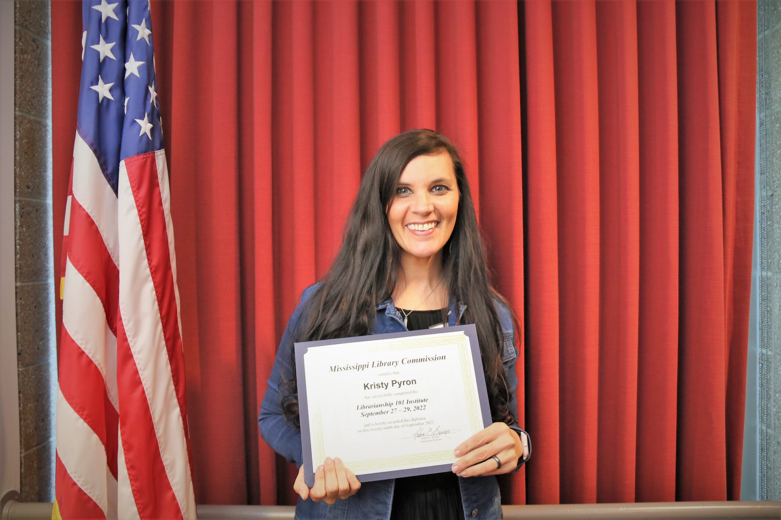 Kristy standing in front of an American flag holding a Mississippi Library Commission certificate.