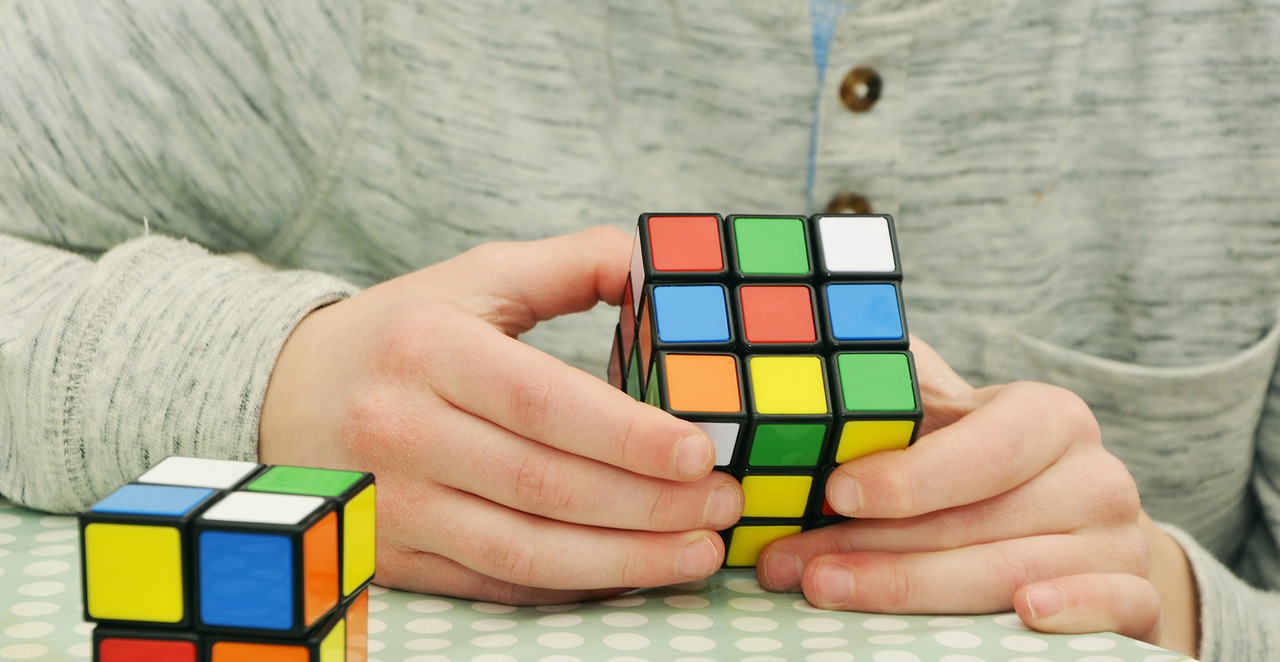 Close-up of hands holding and twisting a colorful Rubik’s Cube, with another cube resting on a table nearby.